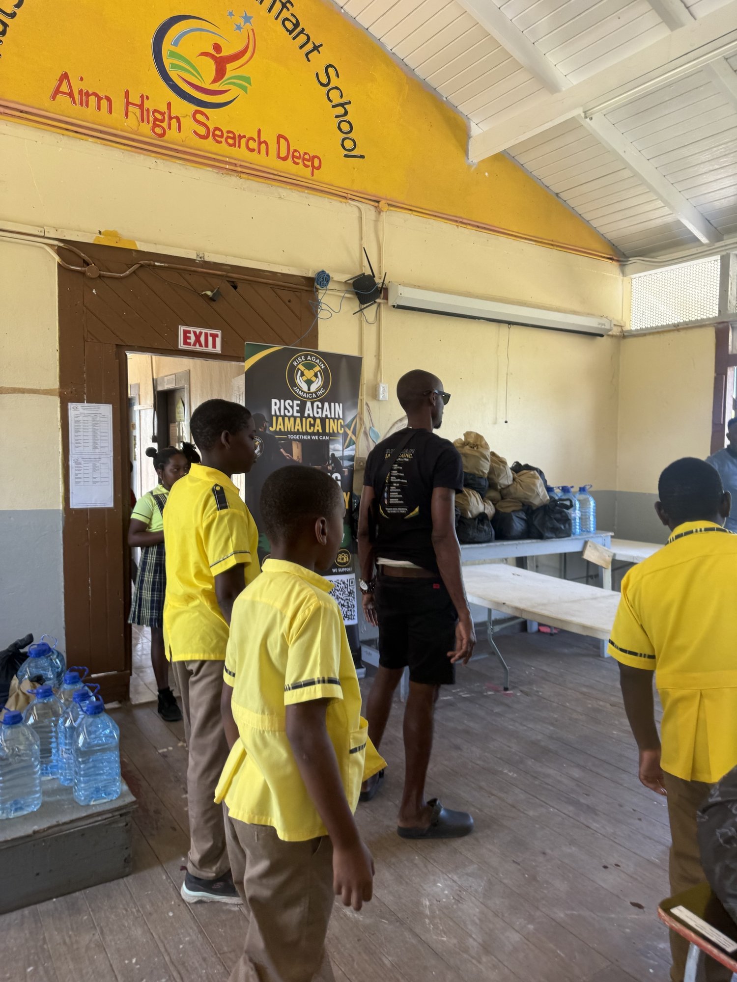 Students in yellow uniforms beside tables of food packages and water at a school distribution.