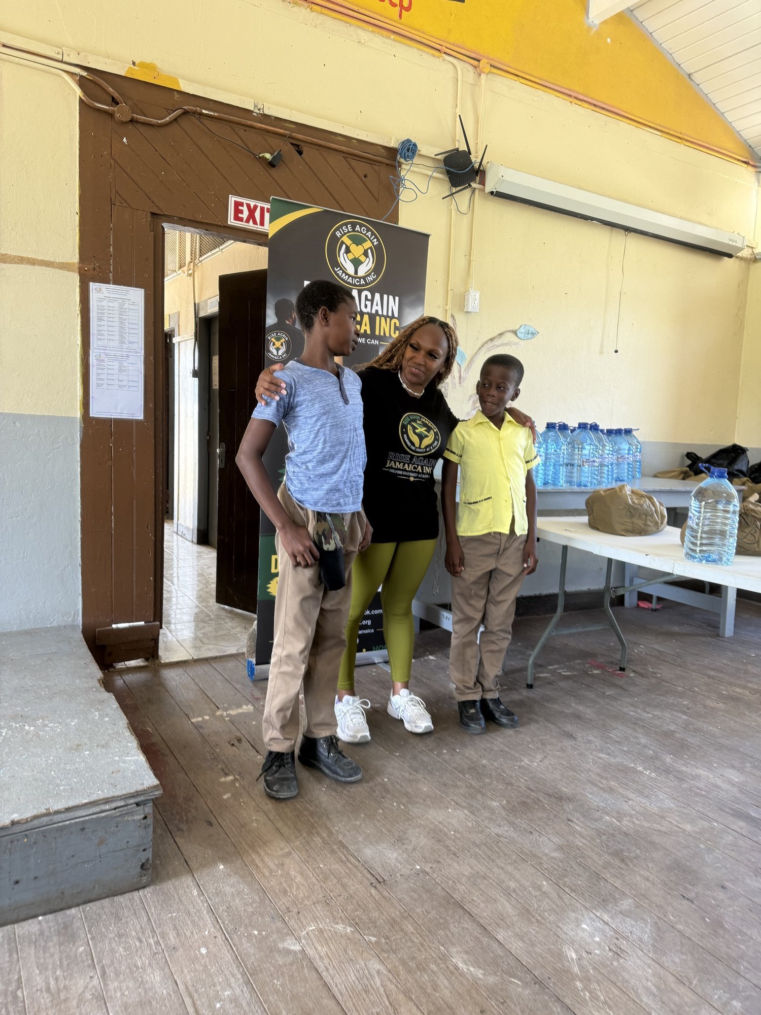 Michelle Ramundo standing with two students in front of the Rise Again Jamaica Incorporated banner.
